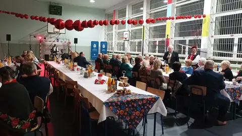 3 zastawione plackami i napojami stoły. Przy ich siedzą seniorzy. Nad stołami wisi efektowana dekoracja w postaci czerwonych korali.