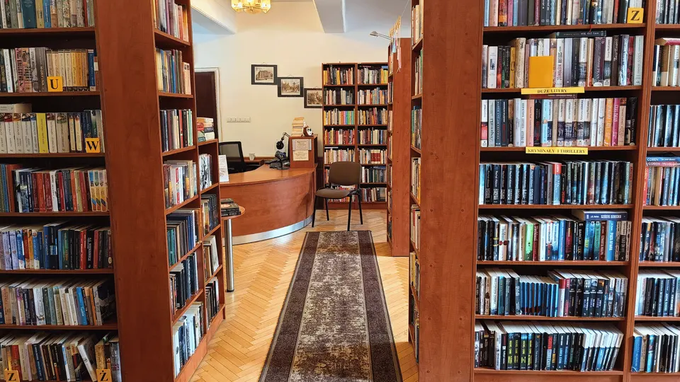 Zdjęcie wnętrza Gminnej Biblioteki w Mogilanach. Po obu stronach przejściach regały książek. Na końcu przejścia stanowisko do obsługi czytelników.