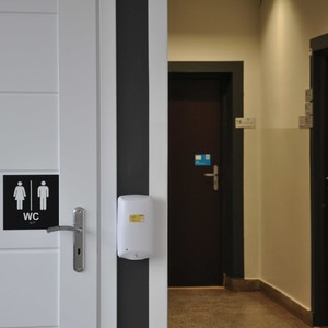 Drzwi ogólnodostępnej toalety oznaczone tabliczką z kontrastowym piktogramem, napisem WC i odwzorowaniem tyflograficznym.