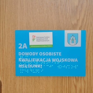 Fragment drzwi do Urzędu Stanu Cywilnego oznaczony kontrastową tabliczką z napisem dowody osobiste, kwalifikacja wojskowa, meldunki wraz z odwzorowaniem tyflograficznym.